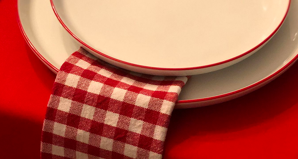 Dos platos de la colección Nordika Red Rim superpuestos, con una servilleta de tela a cuadros rojos y blancos y un mantel rojo
