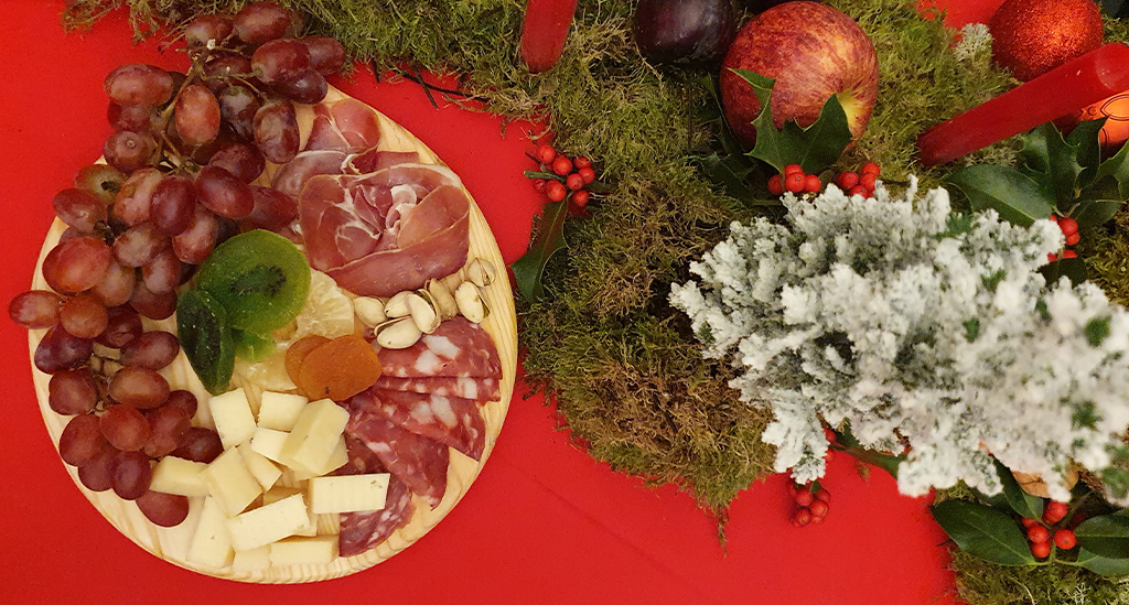 Tabla con quesos, embutidos y varias frutas, a la derecha, y arreglo navideño con plantas y frutas, como manzanas, a la izquierda