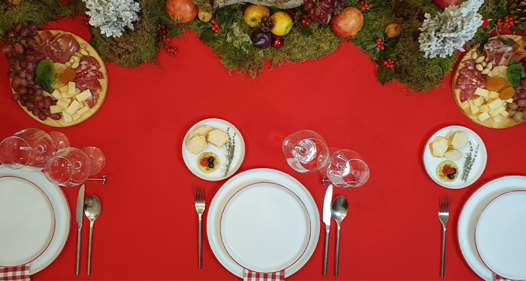 Ejemplo de una mesa para la cena de Navidad con porcelana Nordika Red Rim, centros de mesa de frutas de temporada y mantel rojo