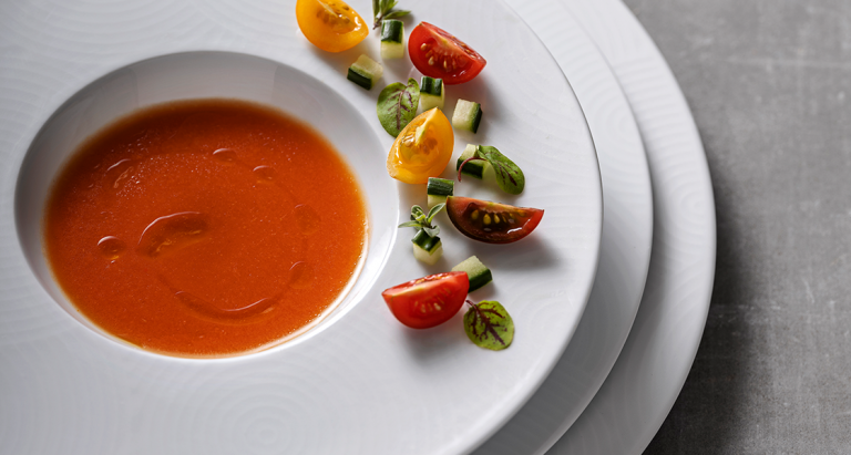 Imagem ilustrativa do artigo “O que é o serviço à francesa” - 3 pratos sobrepostos da coleção Ecos, sendo o prato de cima um prato de sopa com um caldo de tomate e tomates cherry cortados, vermelhos e amarelos acompanhados de ervas aromáticas a decorar a borda do prato.